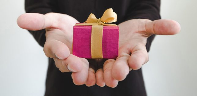 Closeup of a hand holding out a small pink box wrapped with a gold bow as a gift.