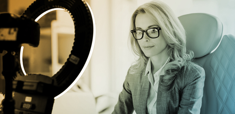 A professionally dressed woman sits at her desk, lit by a ring light while self-recording a video.