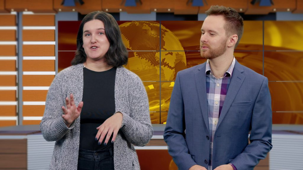 A female and a male speaker in business casual dress present in front of a virtual corporate-looking background.