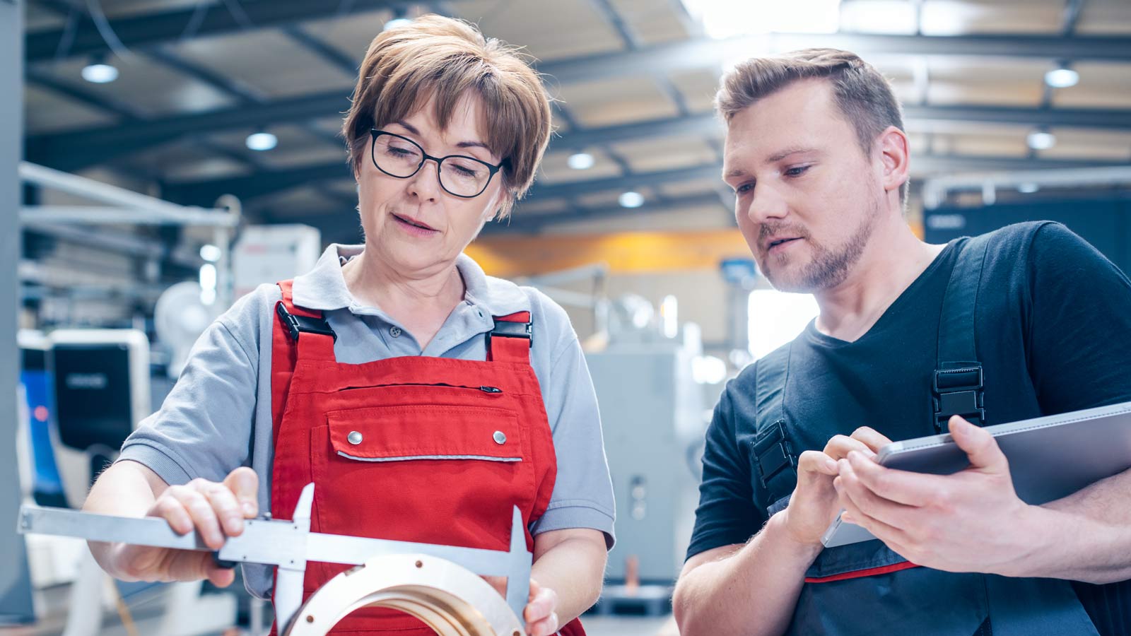 A female factory worker shows her metal work to a male co-worker who is using a tablet computer.