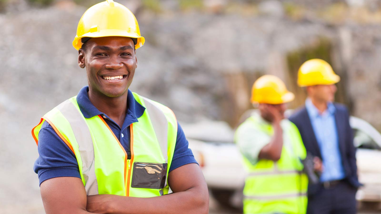 A construction worker wearing a yellow hard hat and safety vest stands in the foreground of a construction site with two additional workers in the background.