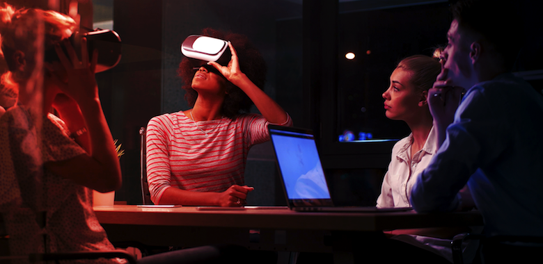 A woman and a man sit with their laptop and observe two other women wearing VR headsets, experiencing virtual reality.