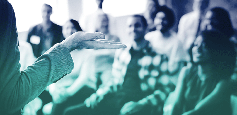 Close up of a female presenter’s hand as she addresses the out-of-focus audience in the background.