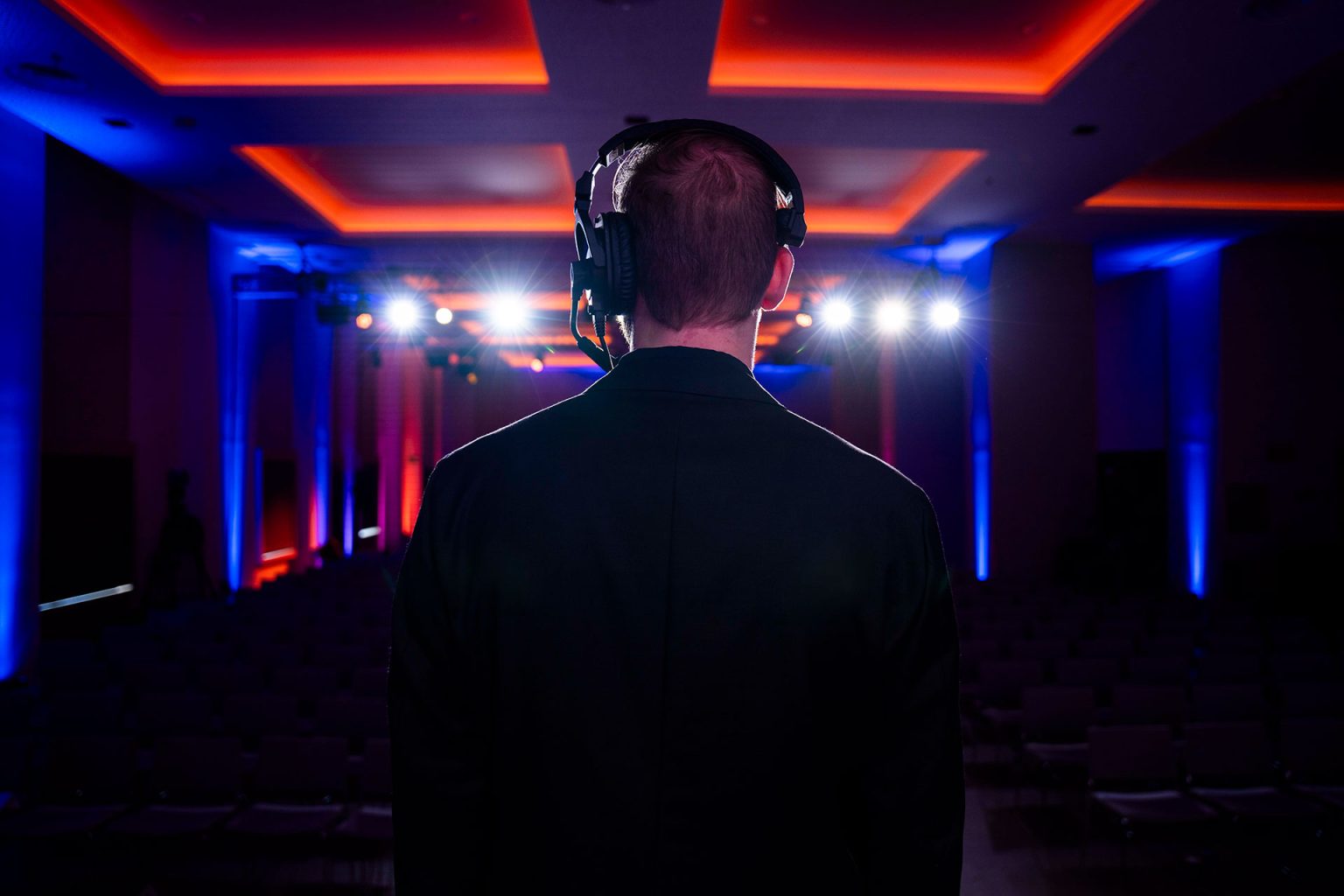 Rear view of a male Brella event management agency producer wearing headset, looking out over darkened corporate event with blue and orange accent lighting.