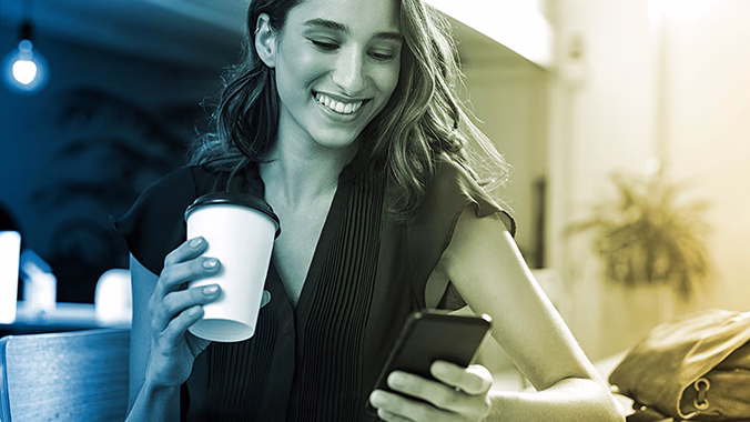 A young woman holding a coffee cup checks her phone.